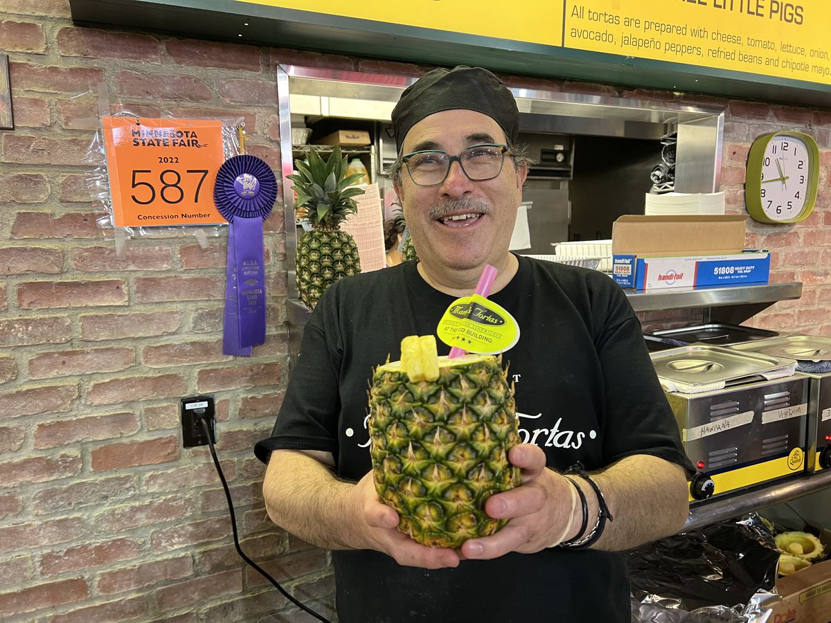 A man wearing a black shirt and black cap smiles and holds a pina colada in a pineapple. 