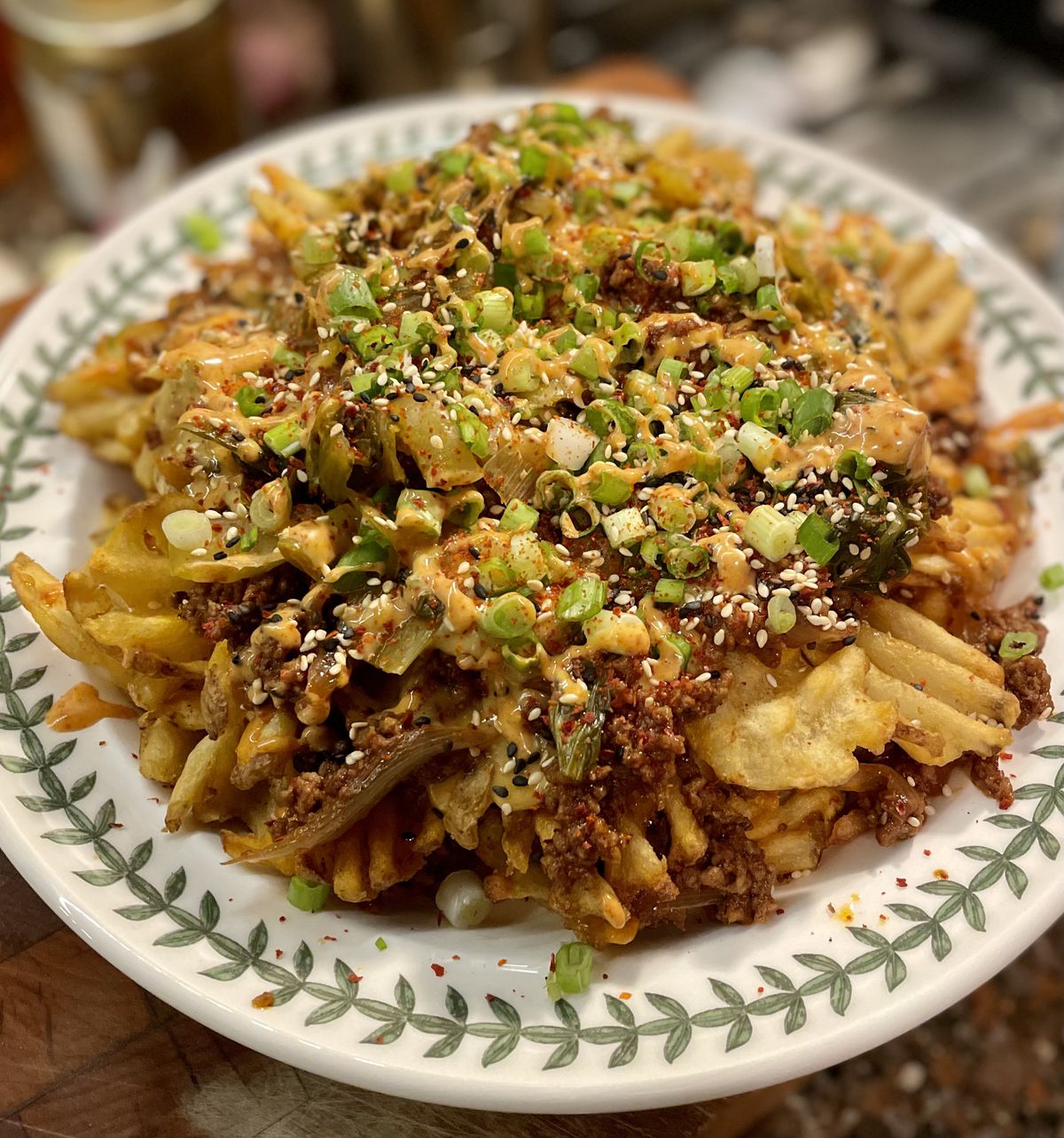 A white and green plate is topped with a mountain of waffle fries topped with bulgogi and kimchi, sesame seeds, and scallions. 