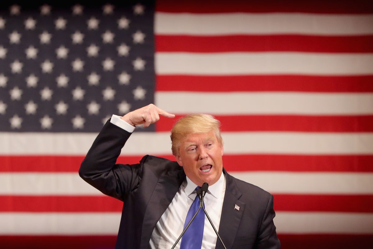 Trump pointing to his own head in front of an American flag.