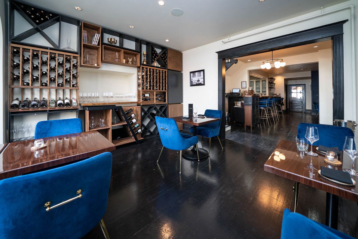 A dining room with a wall covered in a wine rack full of bottles, dark tables, and sea-blue velvet chairs.