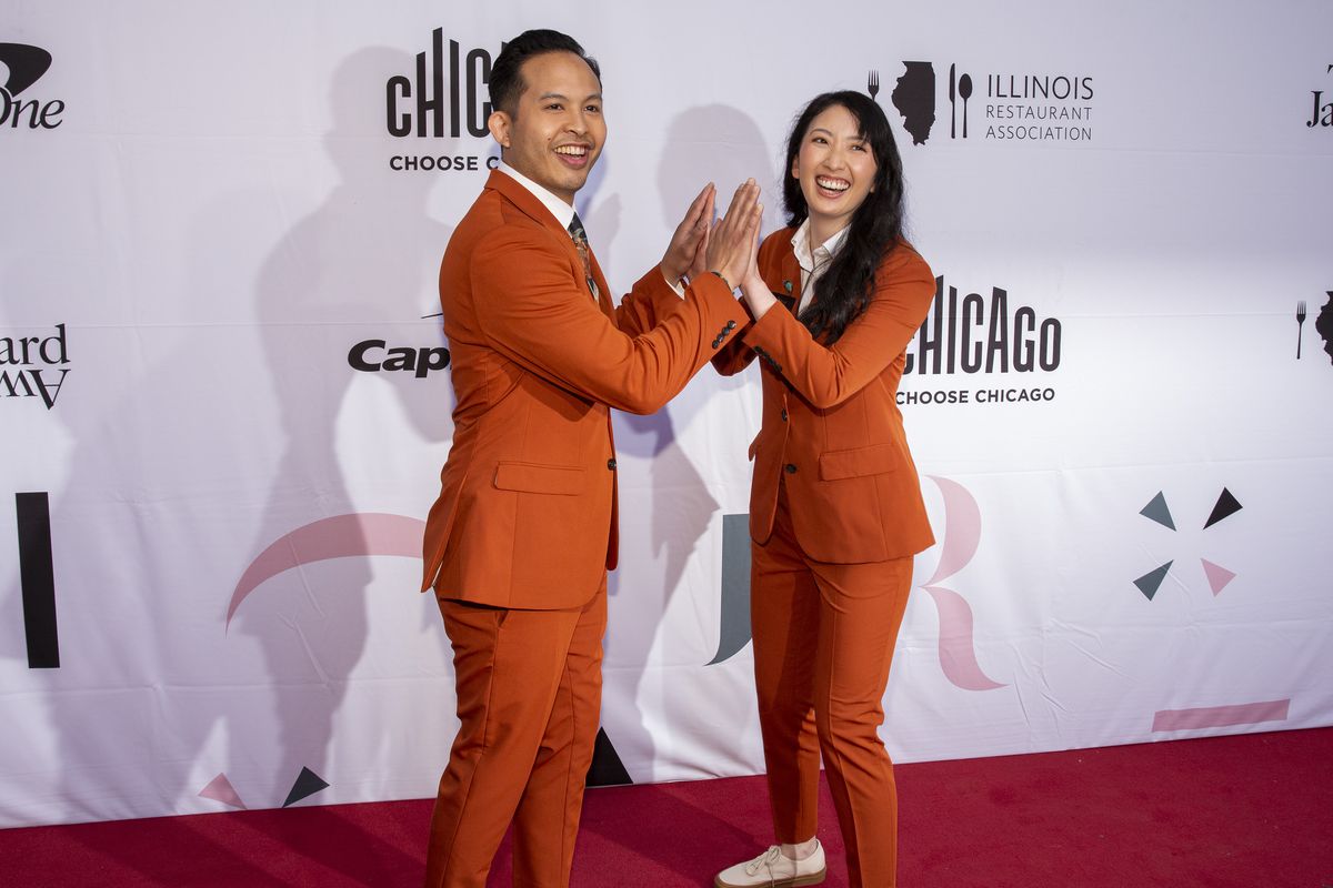 A man and woman in matching red suits pose on a red carpet.