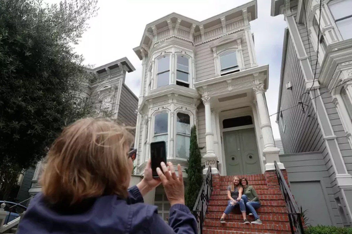 Two people hugging while sitting on the steps of a white Victorian house with red brick steps. A third person in the foreground is taking their picture, her back turned toward us.