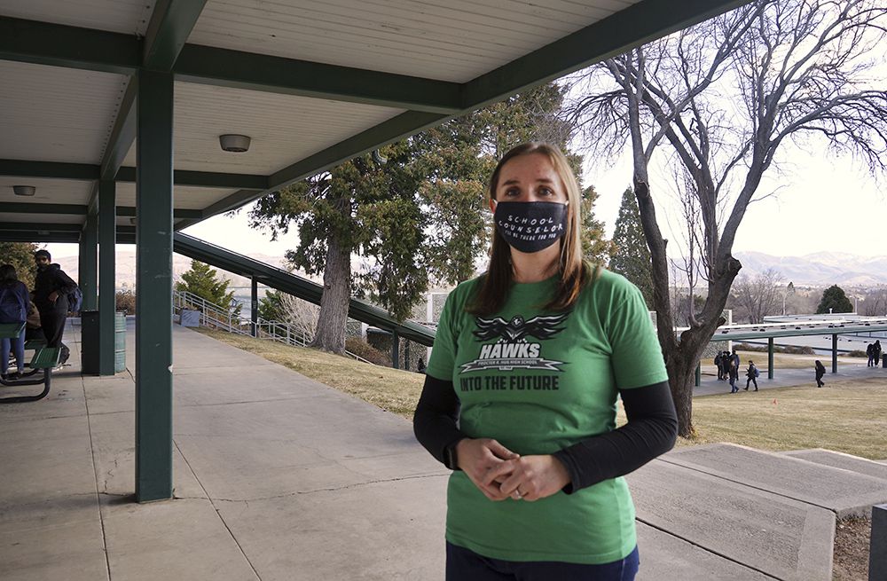 A woman, wearing a black mask and green shirt with the word “HAWKS” in white lettering, stands for a portrait on a high school campus.
