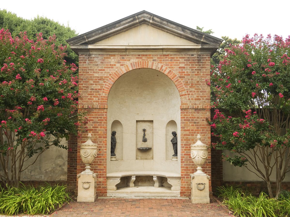 An exterior at the Dumbarton House. The facade is red brick with an archway and altar. There are trees with red flowers surrounding the altar area.