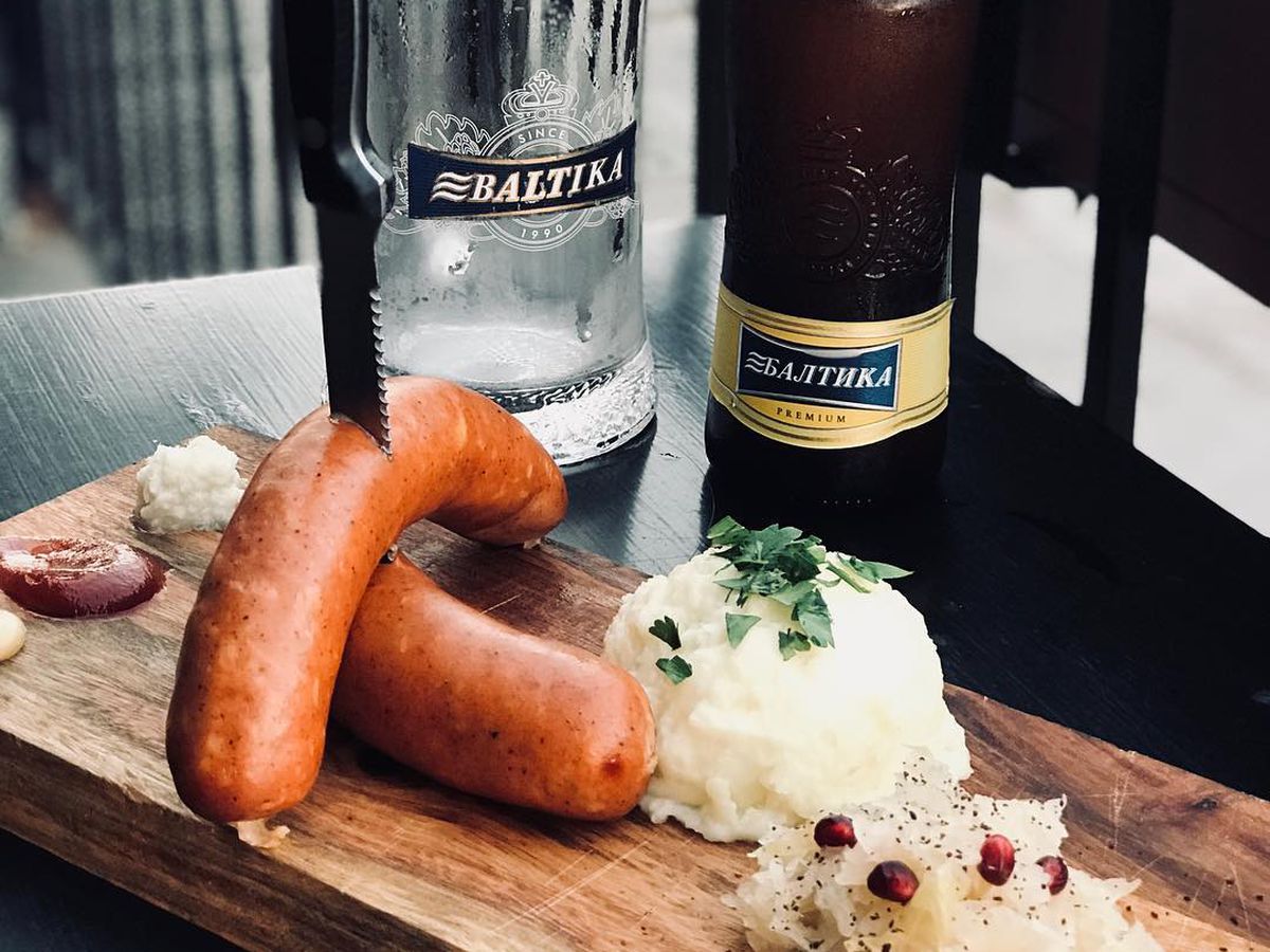 A knife spears two sausages on a board with potato salad and sauerkraut with a beer bottle and pint glass in the background.