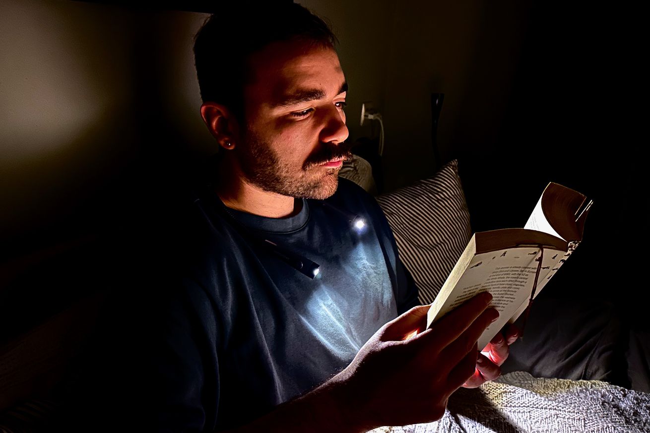 A man in a dark bedroom wears a neck lamp that illuminates just his face and the book he’s holding while sitting upright in bed. 
