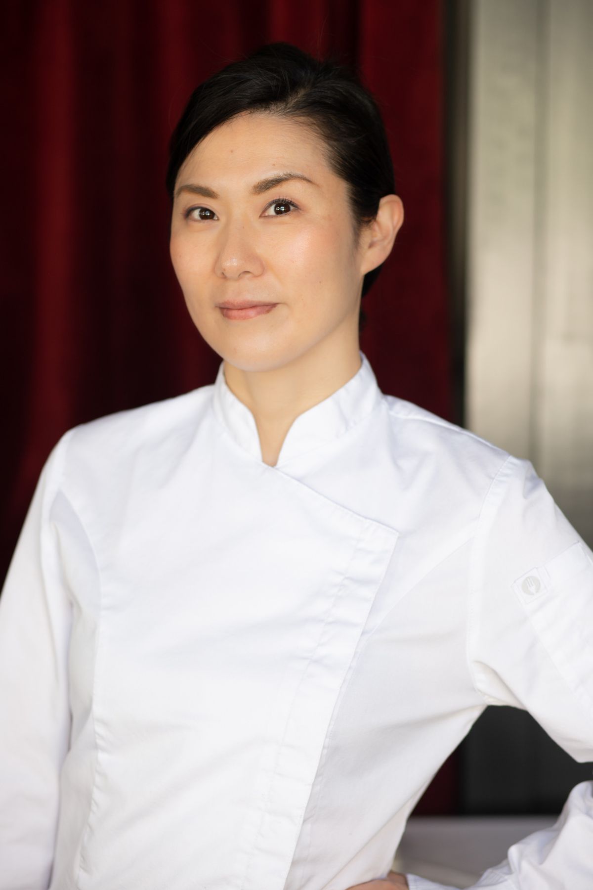 An Asian female chef in a white chef’s coat looks at the camera inside of an empty kitchen.