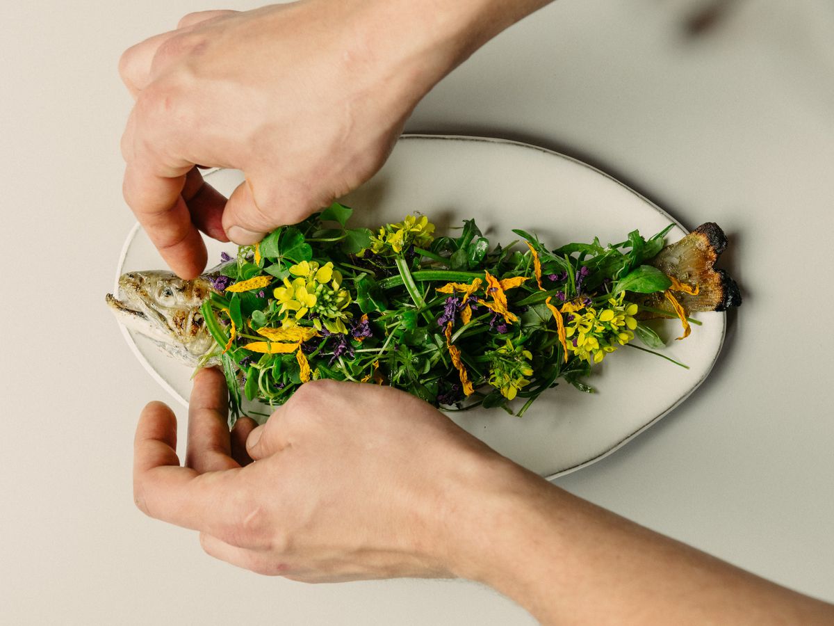 Hands lay a bounty of sprigs and flowers on a whole roasted fish