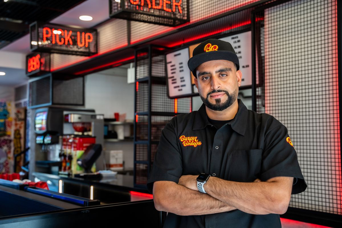 Abbas Dhanani wears a Burger Bodega t-shirt in front of the restaurant counter, which indicates with fluorescent lights where to order, pick up and get food 