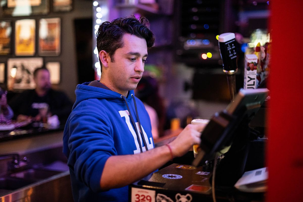 A dark-haired man in a blue sweatshirt works at a pizzeria at night.