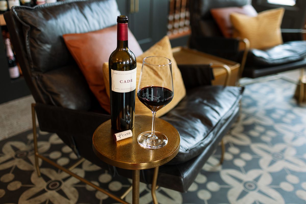 A bottle of wine sits open next to a glass on a small side table in a room full of leather, midcentury furniture.