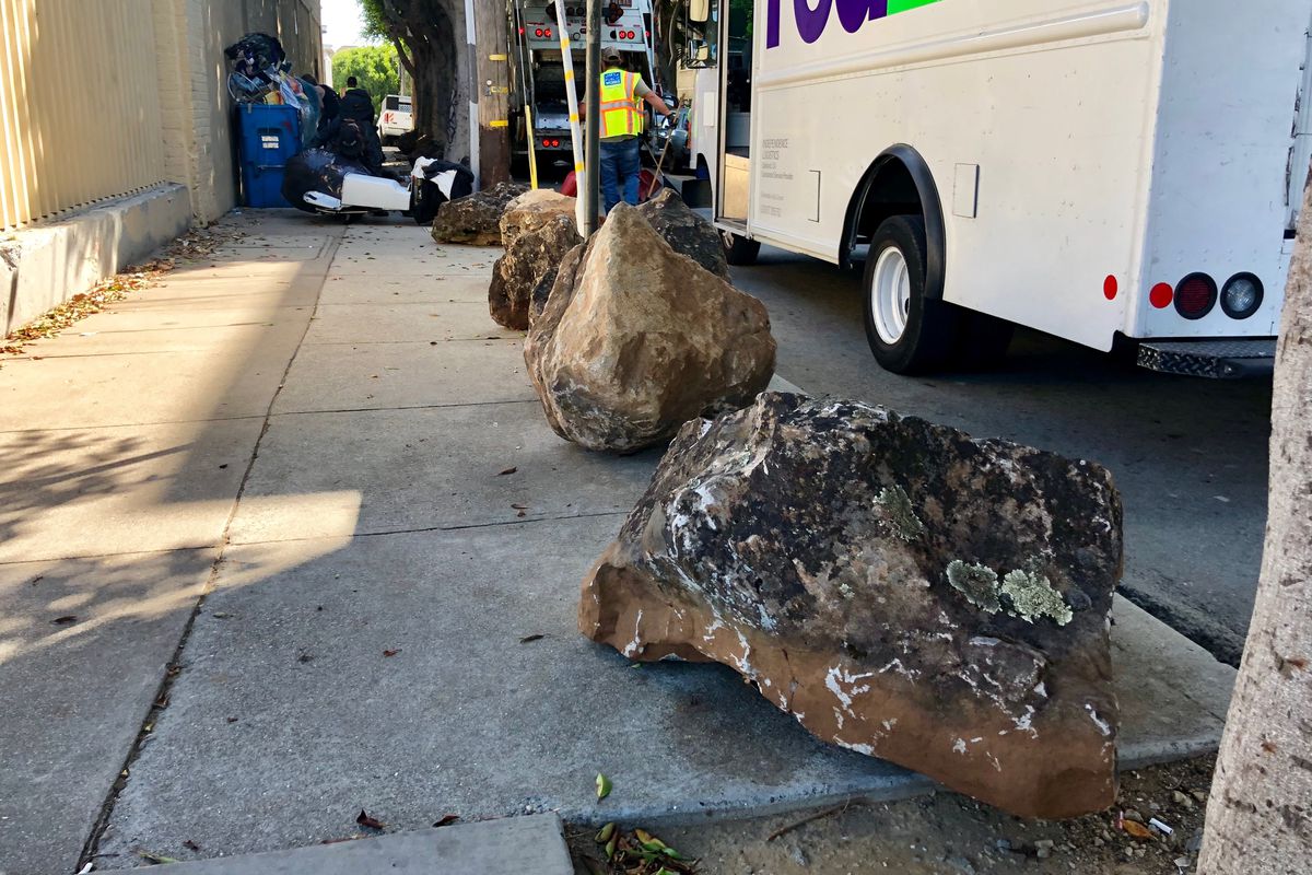 Large boulders lined up along a sidewalk. Homeless people can be seen in the background.