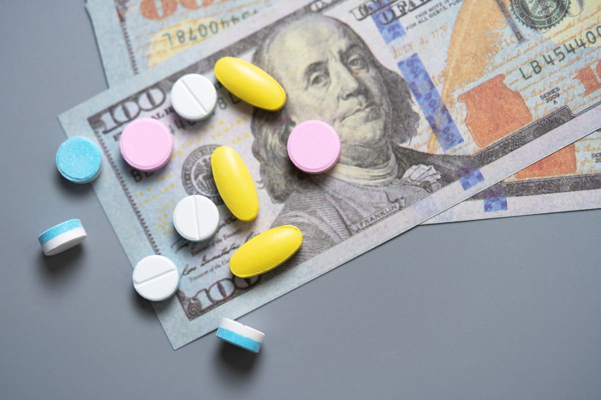 An array of variously colored pills is scattered across a background of $100 bills. 