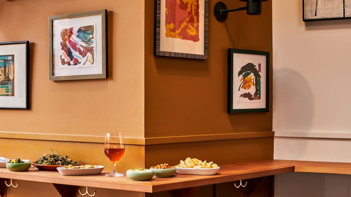 A wooden corner of a wine bar with framed art, tan walls, and light red wine surrounded by snacks.