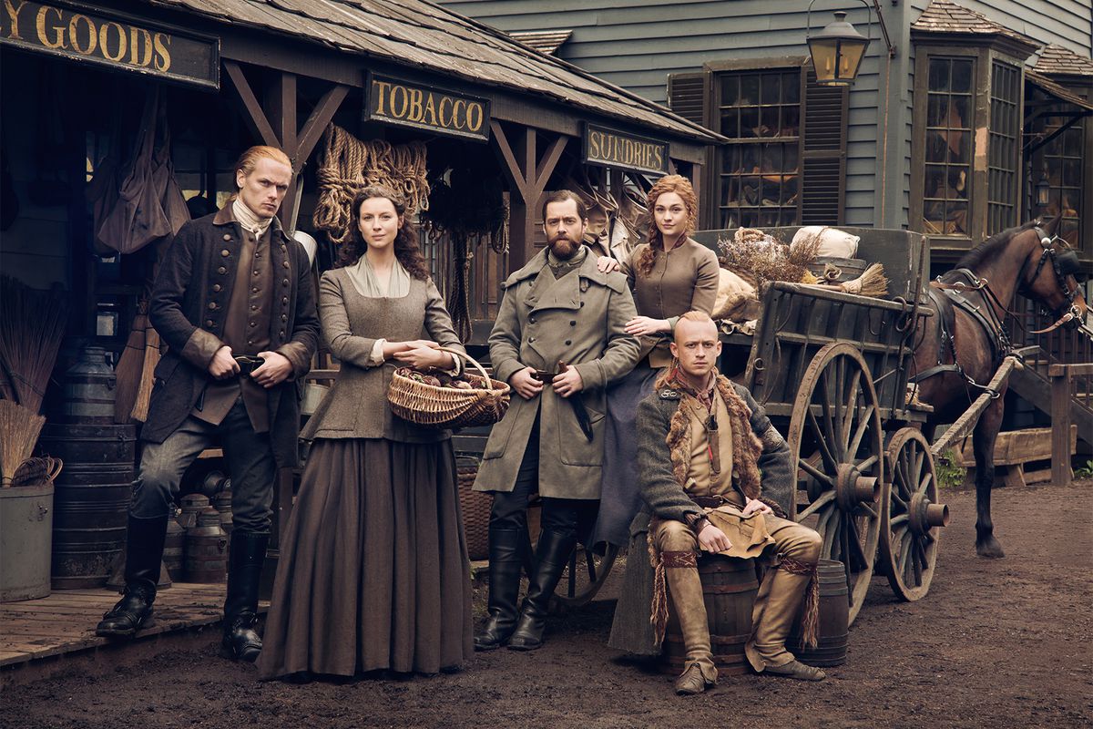 brianna randall (sophie skelton), Claire Fraser (Caitriona Balfe), exterior, gallery, jamie fraser (sam heughan), roger wakefield (richard rankin), young ian (john bell) stand in front of an old timey town square in Outlander season 6