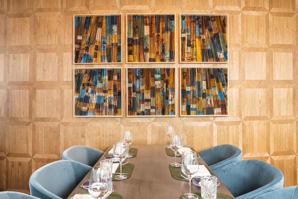 A set table with blue chairs is against a wooden wall. Above it are six pieces of artwork made from folded paper.