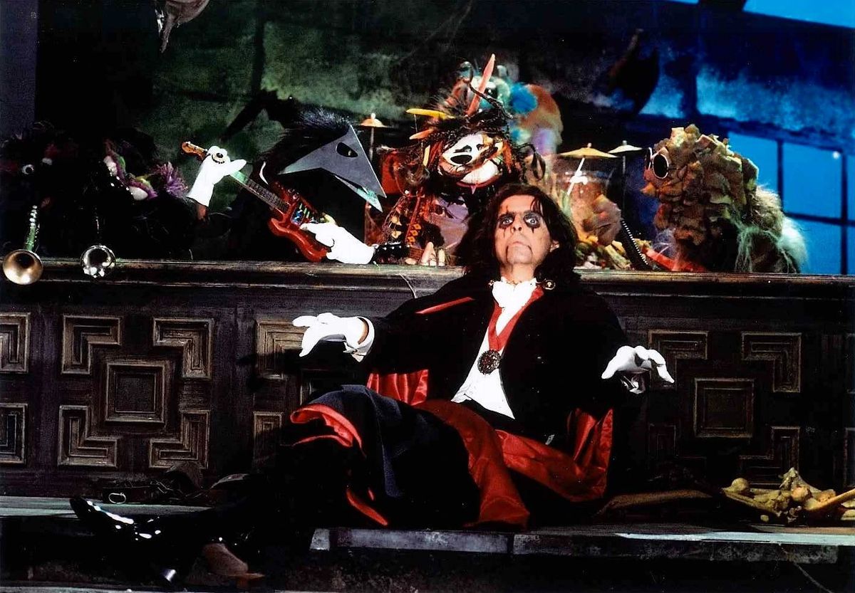 Alice Cooper, wearing bleeding eye liner and dressed in a vampire costume and cloak reminiscent of Bela Lugosi’s Dracula, sits in front of a band of puppet players.