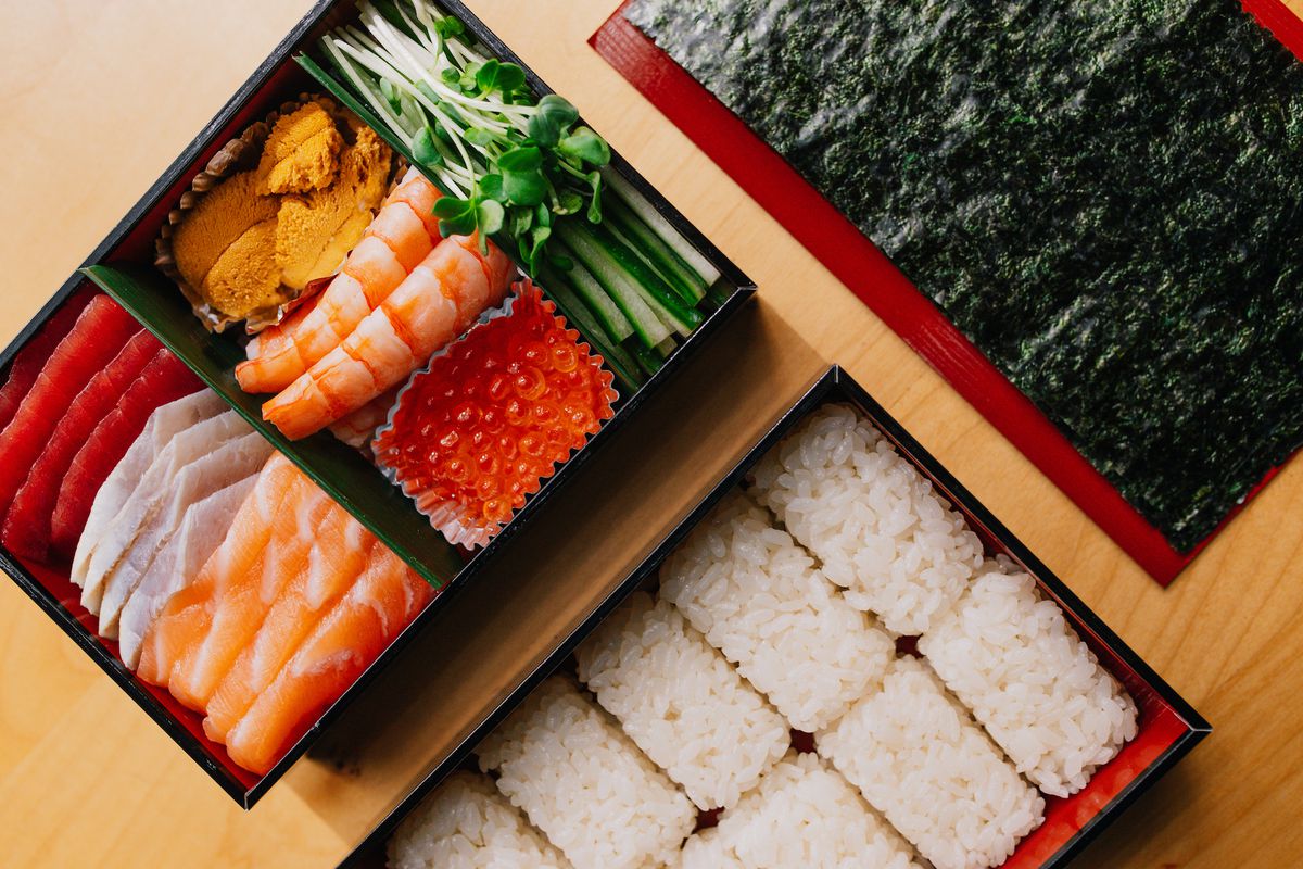 From above, a shot of sushi takeout in a box, with nori and rice on the side.