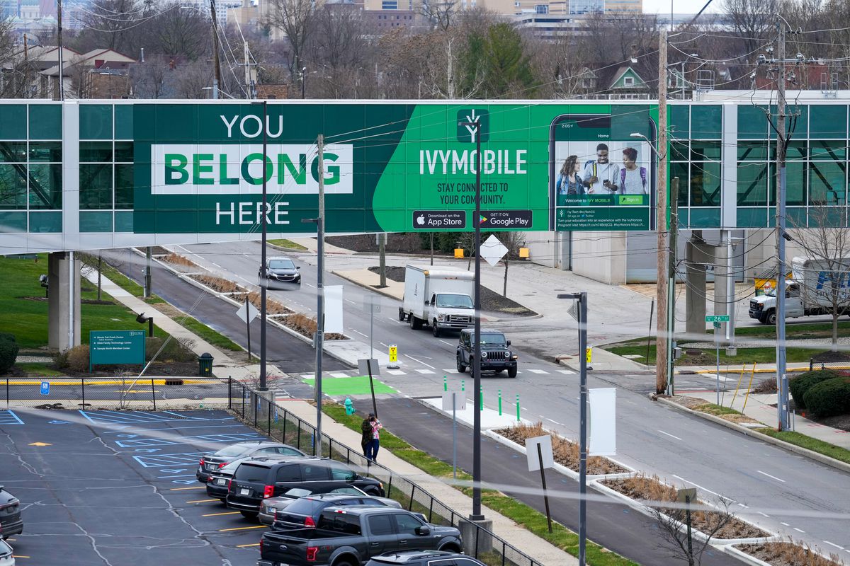 A green sign on a building’s connecting bridge reads “YOU BELONG HERE”, and advertises Indiana’s Ivy Tech Community College. Under the bridge, cars drive down a street near a college campus.