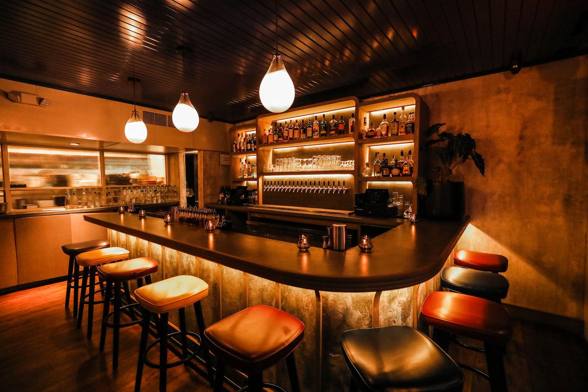 A dimly-lit bar with large lightbulbs above, a dark brown bar, and orange leather covered seats behind it.