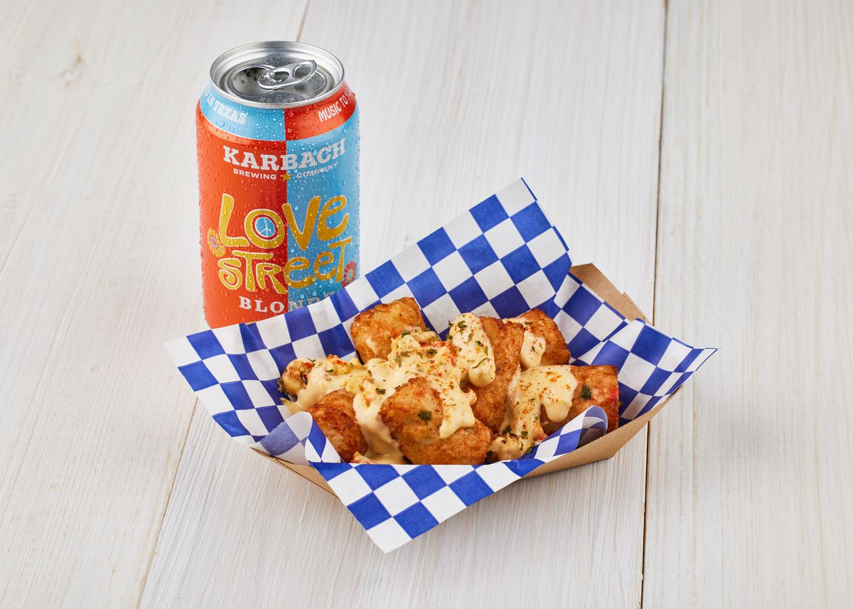 A boat holds tater tots covered in an orange-tinged sauce. A blue and orange can of beer sits behind.