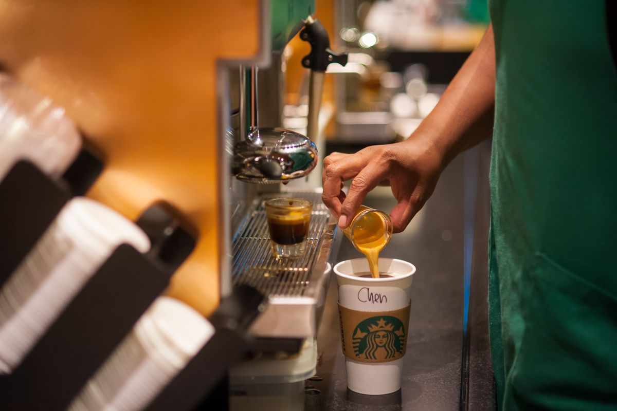 A closeup of a Starbucks employee’s hand pouring espresso into a paper to-go cup with Starbucks branding.