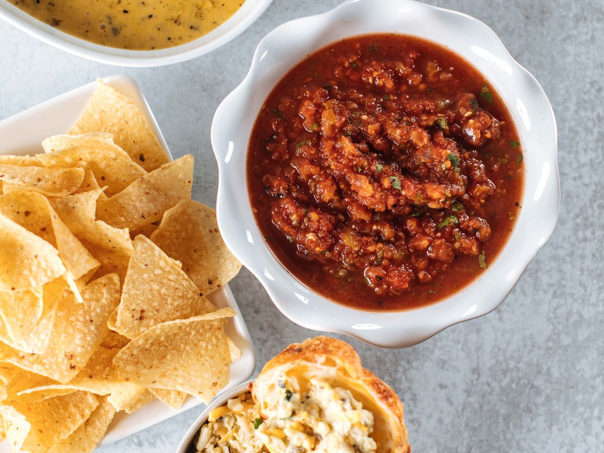 A trio of dips, including salsa, pimento cheese, and queso, sit on a white table next to a bowl of tortilla chips.