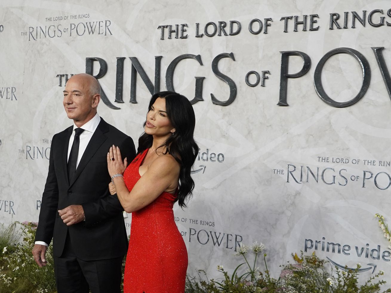 Jeff Bezos, in a black suit, and Lauren Sanchez, in a red dress, are being photographed standing in front of a backdrop reading “The Lord of the Rings: Rings of Power.”