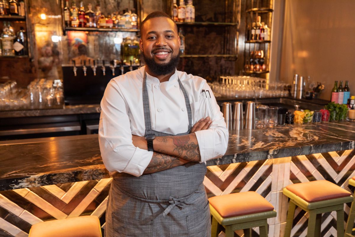 A Black male chef in front of a restaurant bar.