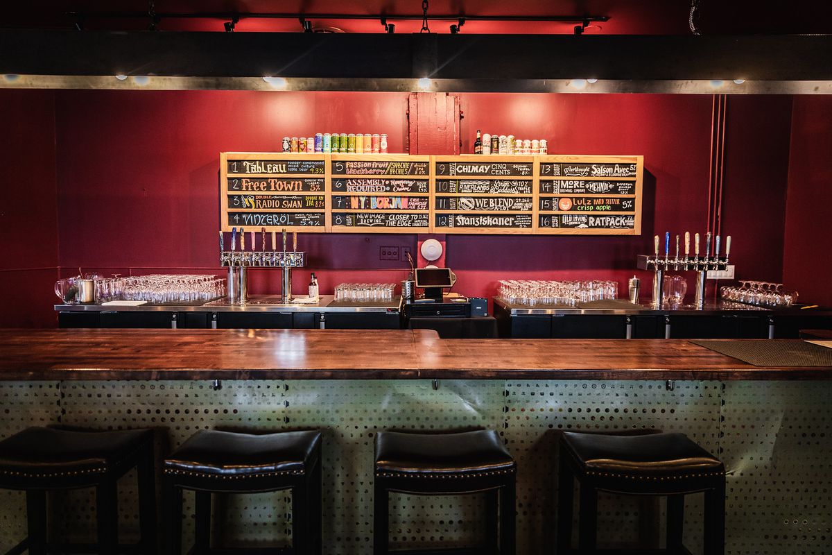 A bar with a wood top and metal base with backless stools in front. Behind, two rows of taps, glasses, and  a chalkboard menu with cans of beer on top.