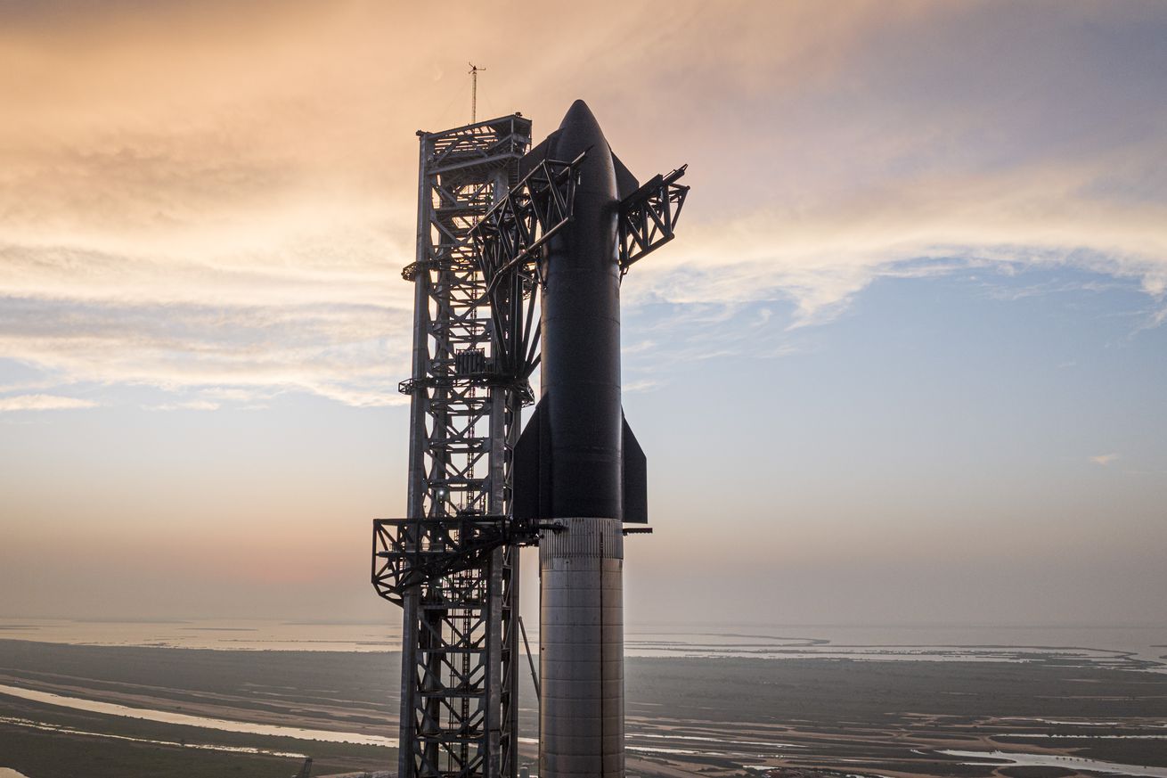 SpaceX Starship prototype stacked on top of a Super Heavy rocket in Boca Chica, Texas, mounted on a launch tower shown with a sunset and partially cloudy backdrop.