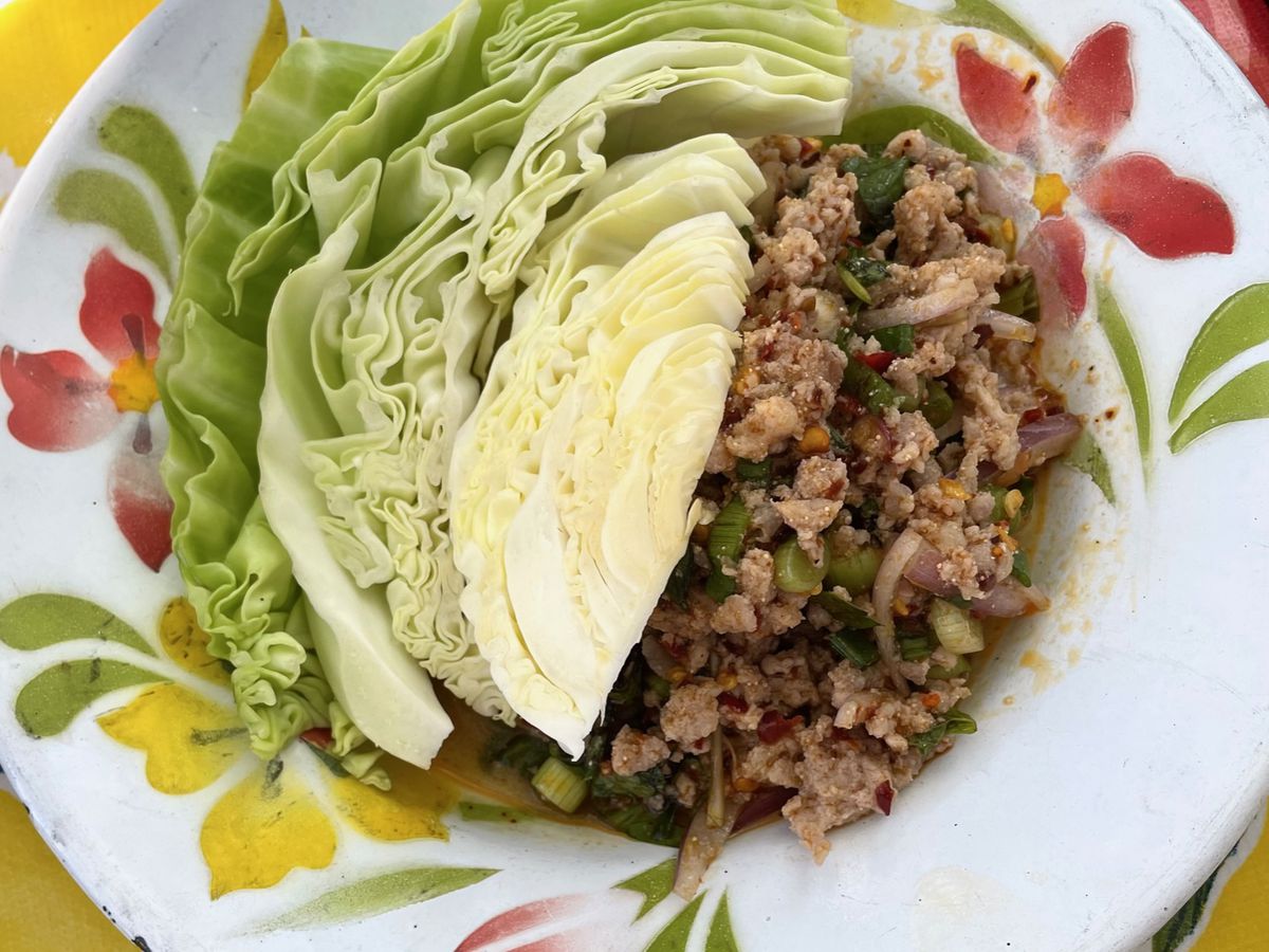 A plate of Thai minced pork (larb moo) dotted with toasted rice sits next to a wedge of fresh green cabbage on a white plate painted with maroon and yellow flowers.