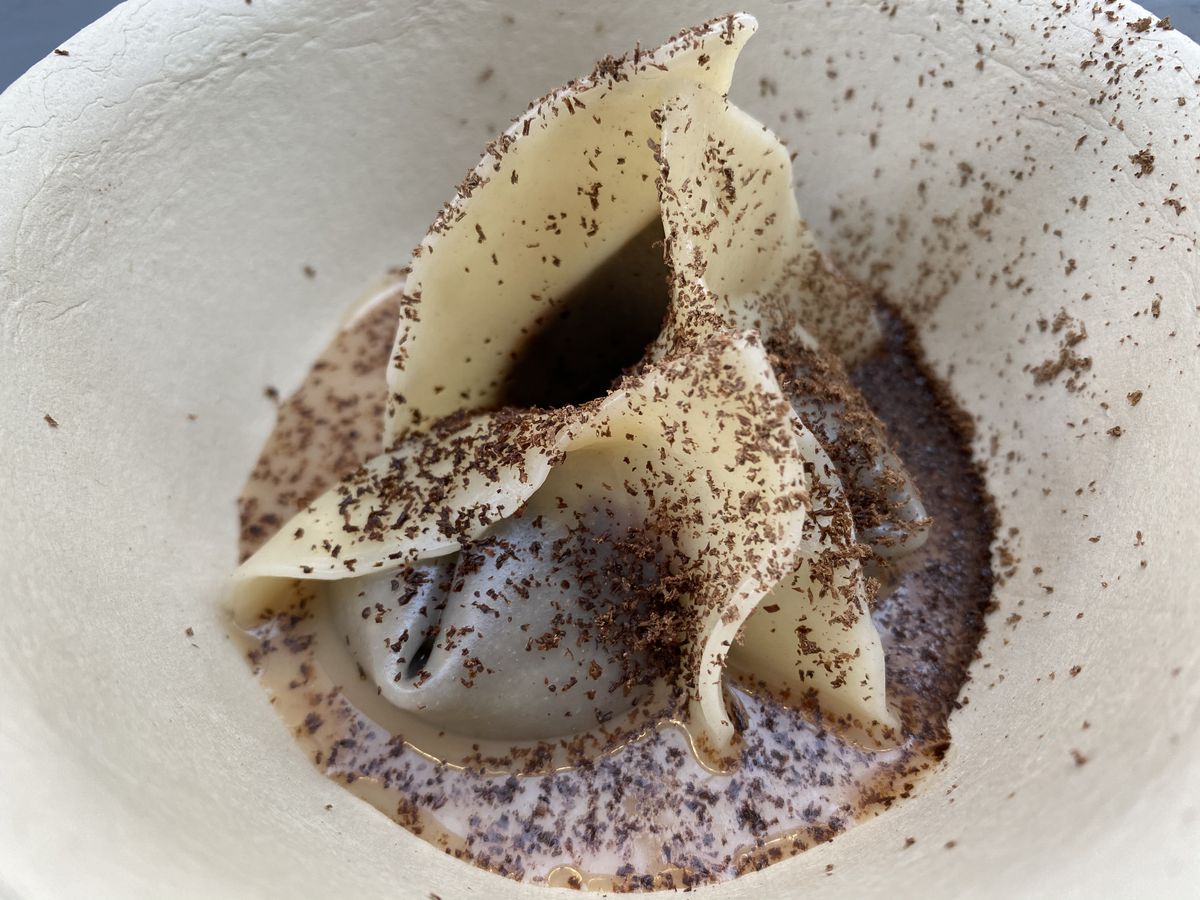A dessert dumpling served with dark chocolate ganache inside sitting in a small pool of milky tea in a tan compostable serving bowl