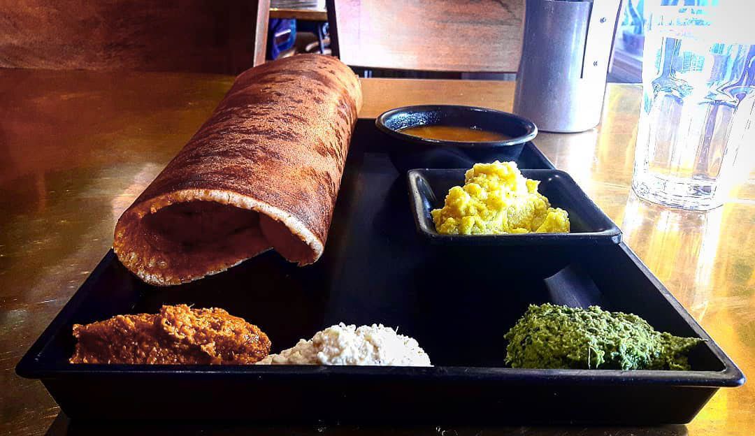 A collection of food items on a tray including a large rolled dosa flatbread and individual small plates of dips.