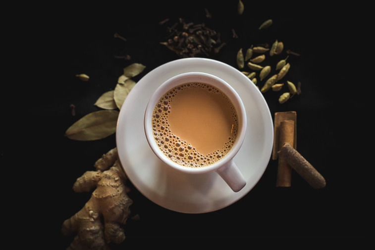 A white mug of rich brown chai on a black background with some spices. 