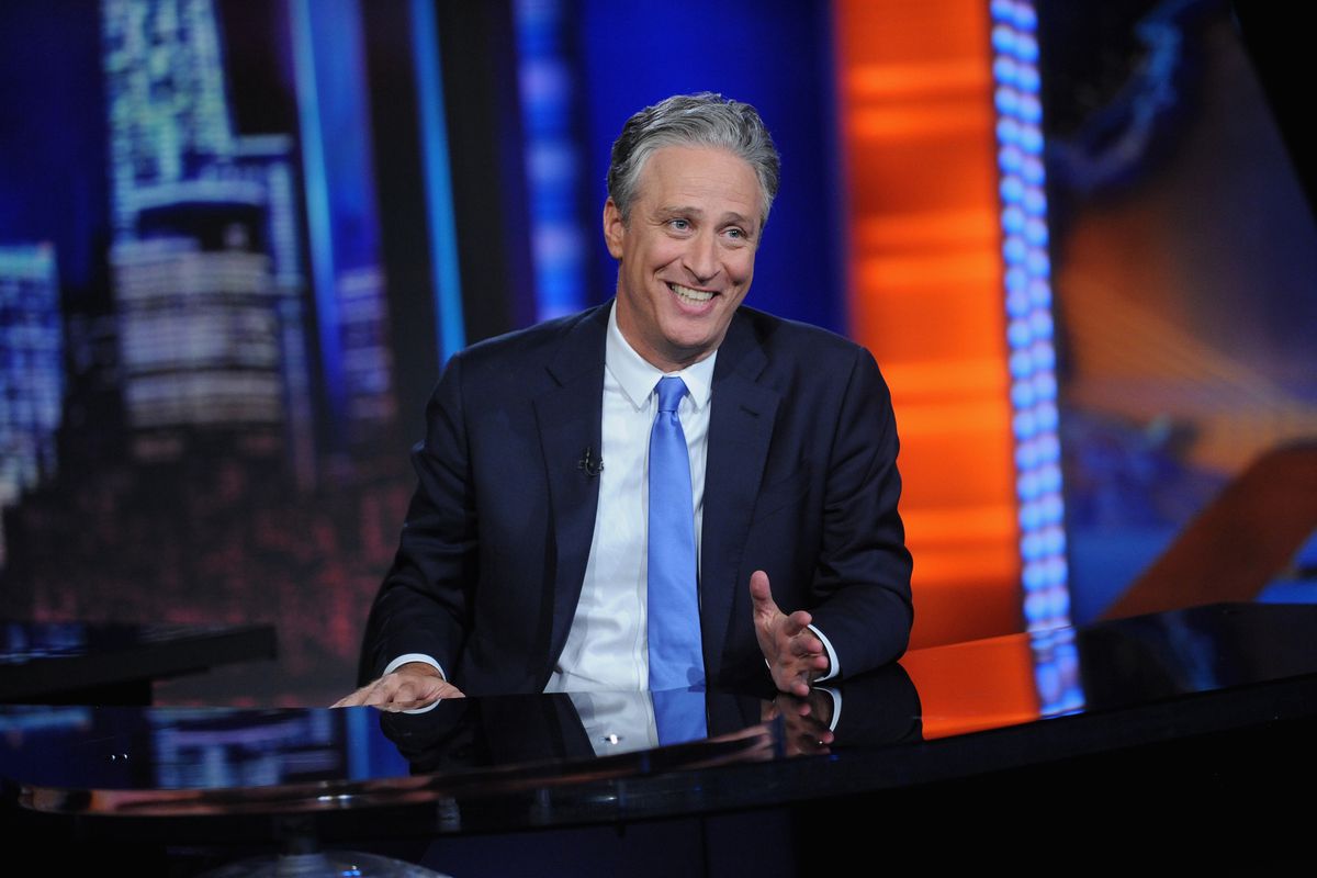Jon Stewart sits at a desk wearing a black suit jacket and light blue tie, smiling. 