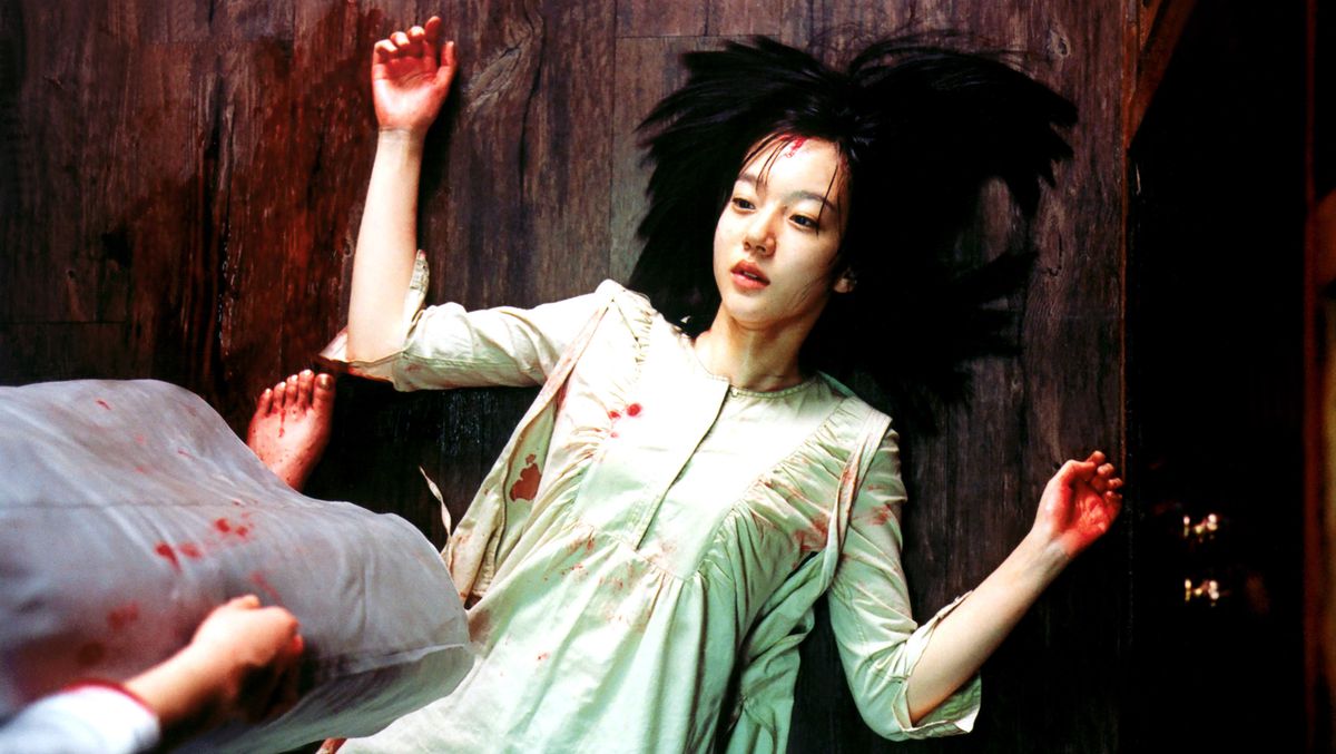 A Korean woman with bloody hands and spots of blood on her dress lies on her back on the floor, hair spread around her in a halo, as a figure with bare, bloody feet stands over her in A Tale of Two Sisters
