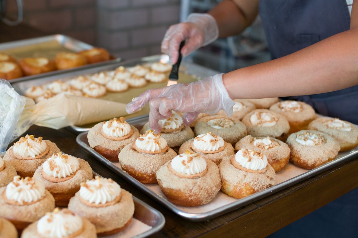 A person’s hands are decorating rows of doughnuts with white frosting lined up in a sheet pan