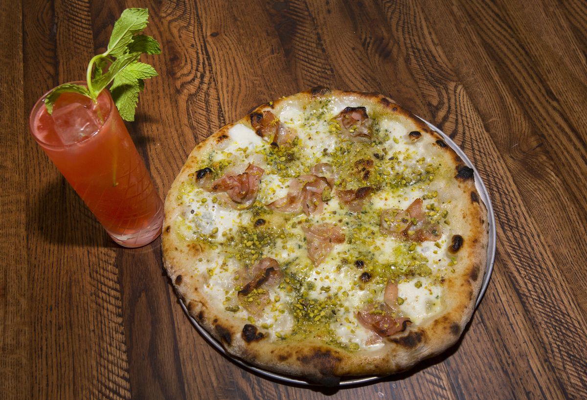 A pink cocktail in a tall glass with herbs sticking out next to a thin-crust pizza.
