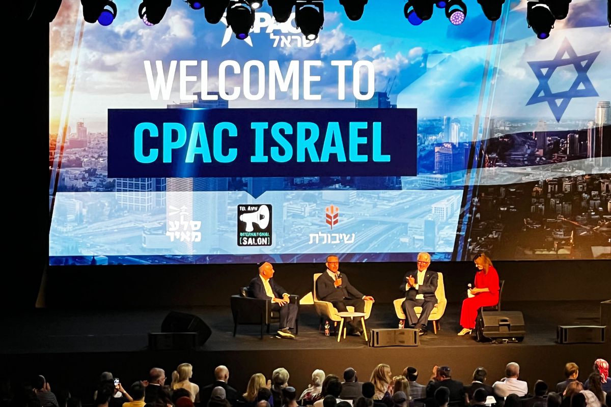 Four people sit on a stage. Behind them a large screen reads “Welcome to CPAC Israel.” 