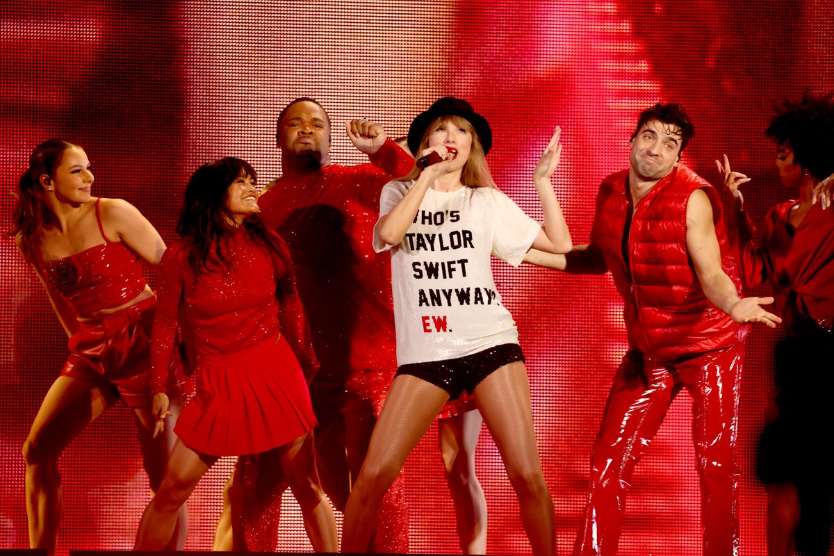 A singer in a white shirt that reads Who Is Taylor Swift Anyway? Ew. With dancers in red behind her.