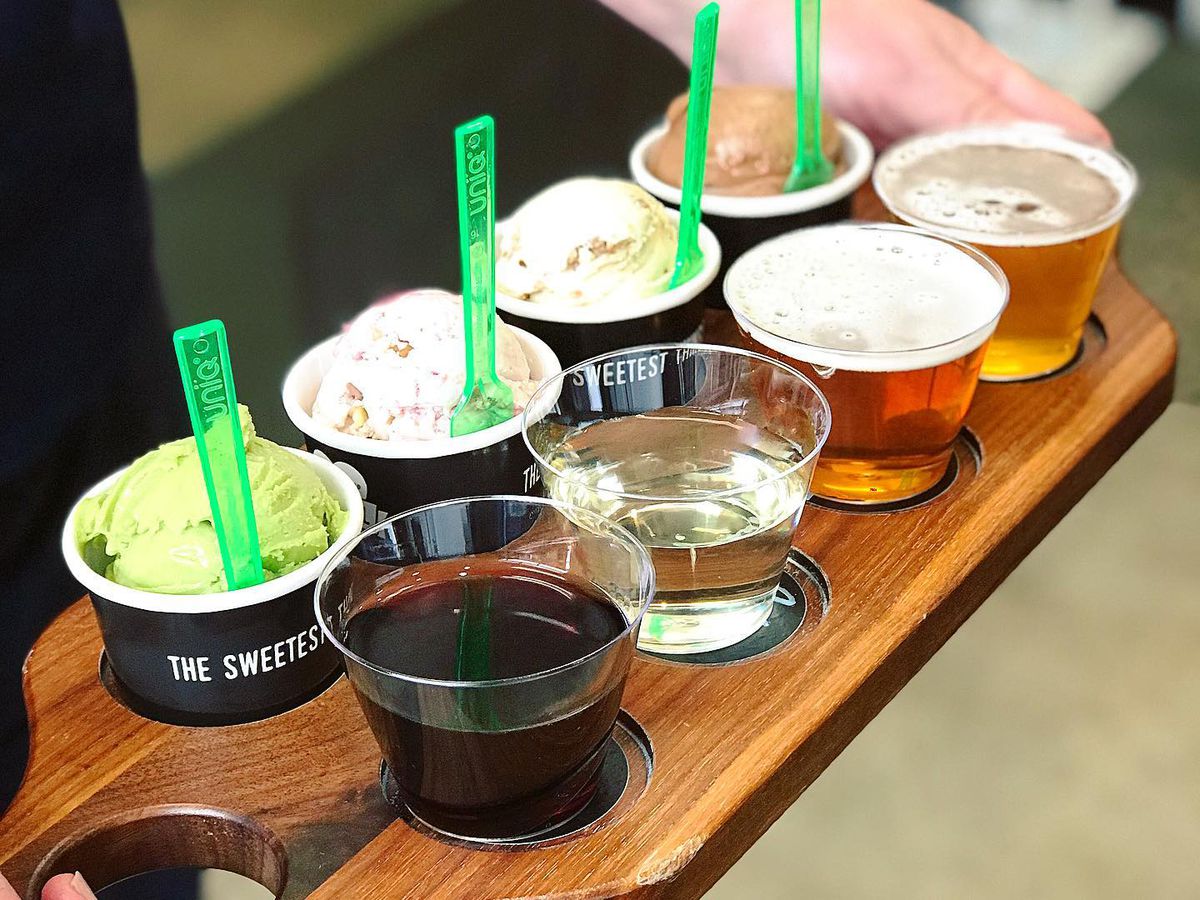 A server holds a wooden serving board with cups of ice cream and glasses of drinks inserted into divots. 