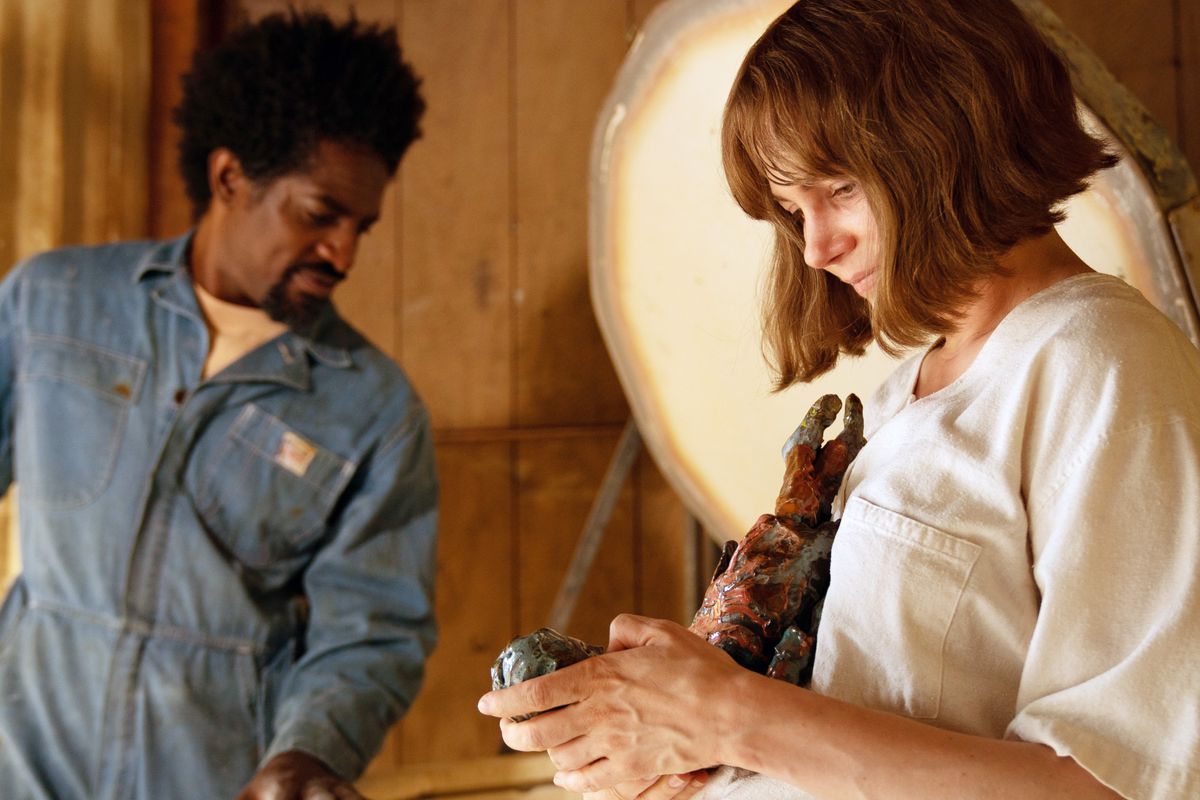 Lizzy (Michelle Williams) smiles over one of her clay sculptures as Andre 3000, in denim coveralls, stands in the background in Showing Up