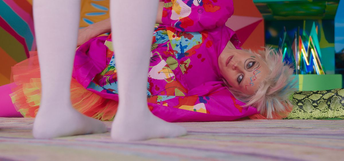 Weird Barbie (Kate McKinnon), a Barbie in a shapeless, baggy, multicolored dress, with her hair cut at various short lengths dyed pastel pink and blue, and with scribbles on her face, lies on the ground staring at the stockinged, shoeless feet of Barbie (Margot Robbie) in the 2023 live-action movie Barbie.
