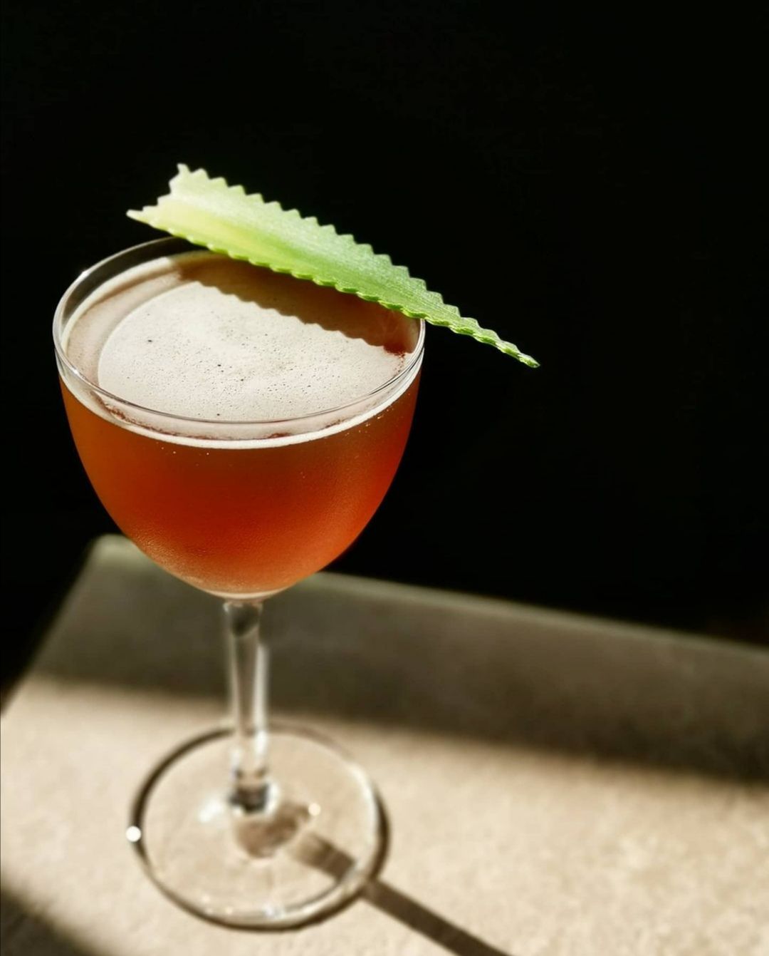 An orange-brown cocktail in a delicate glass is garnished with a green leaf with ridged edges. The drink sits on a stone bar.