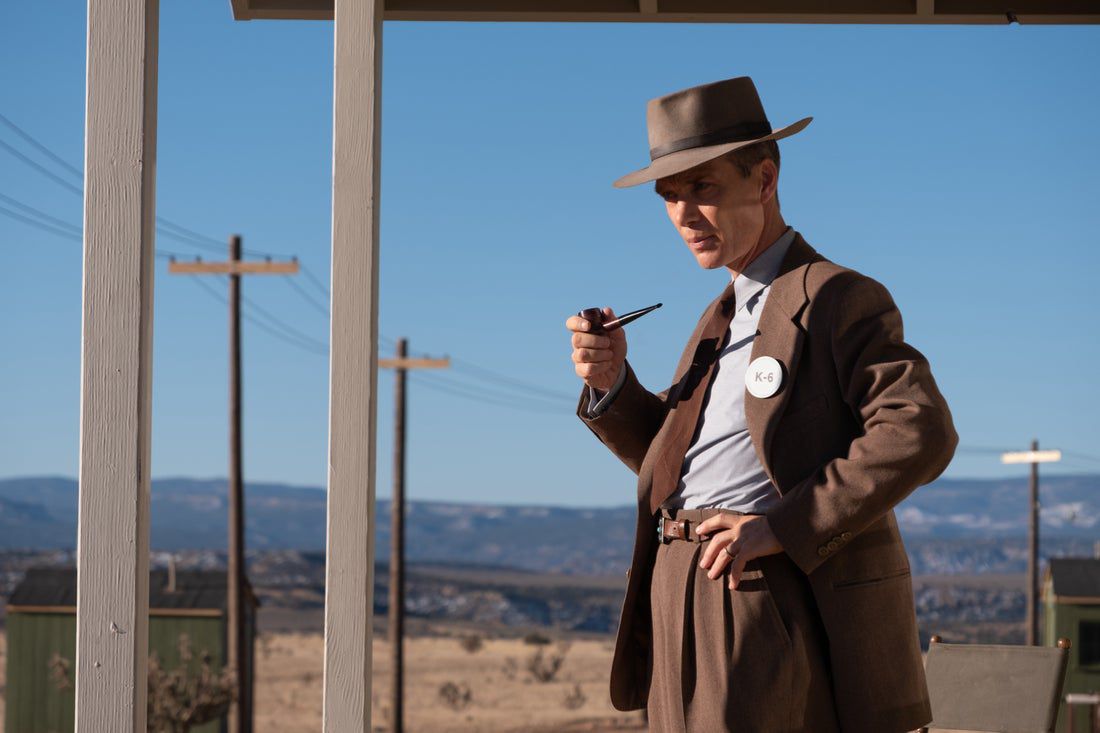 A man in a wide brimmed hat and suit smokes a pipe in a desert with phone lines behind him.