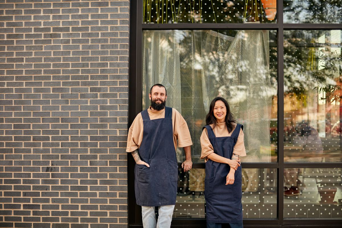 A man and woman in matching blue aprons pose and smile outside a restaurant space.