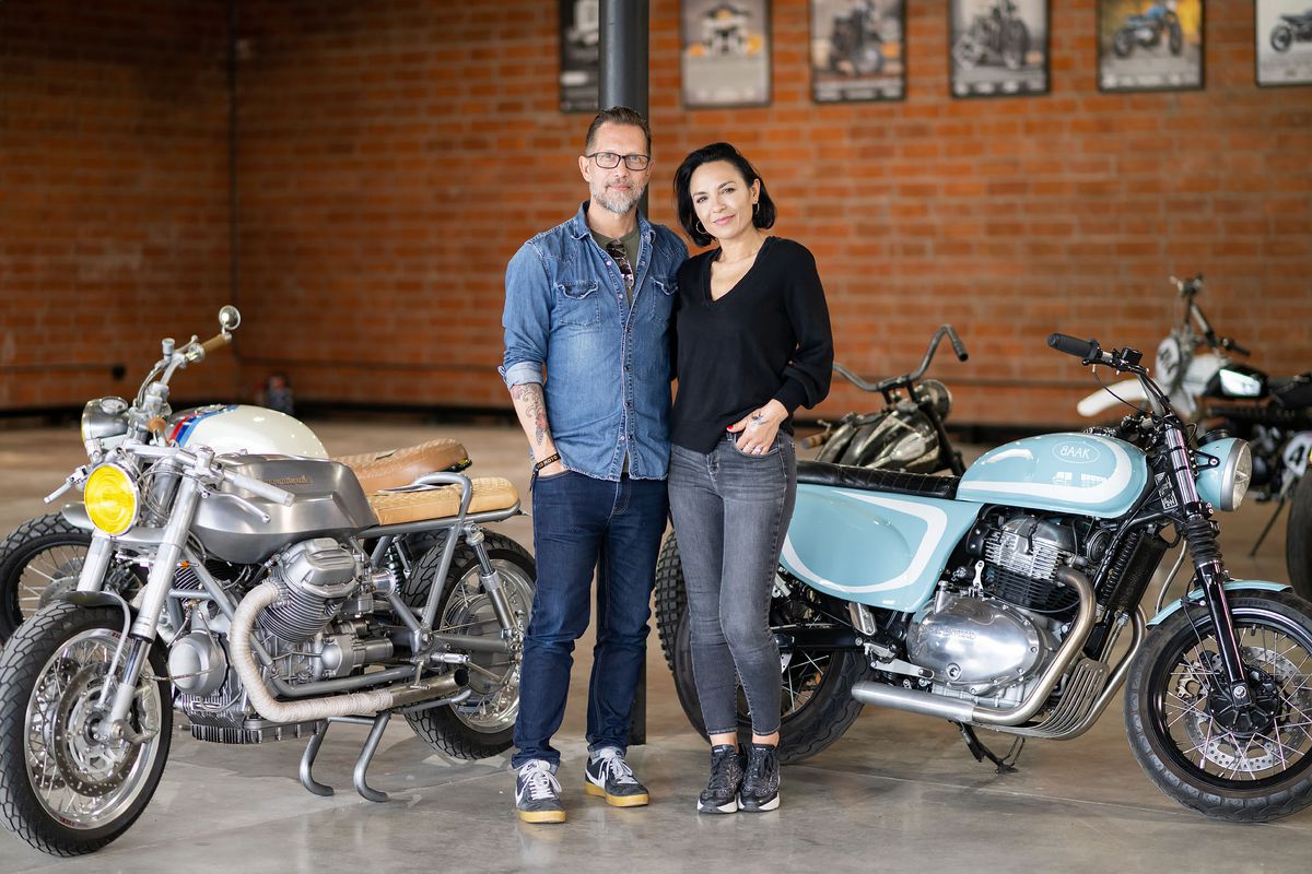 Two people in casual clothing stand next to vintage motorcycles inside a brick building.