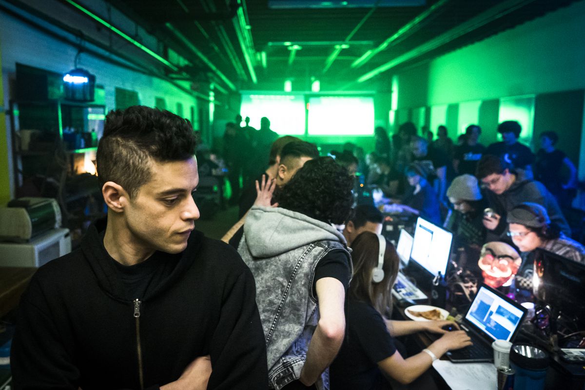 A scene in a dark room filled with people at computers and standing around, with a green light in the background and a young man in the foreground.