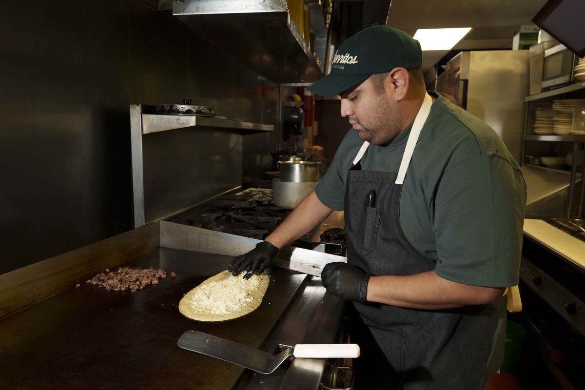 Miguel Hernandez, wearing a black apron, T-shirt, and Litos Burritos hat, putting cheese on a tortilla on a flat-top grill.